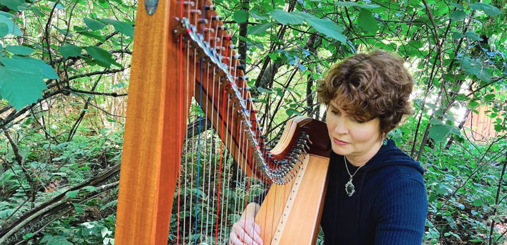 Jen Midkiff sits at the harp with lush green woods in the background.